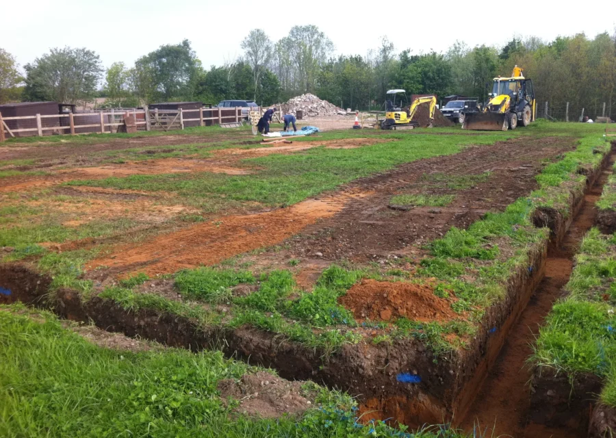 Open drainage trenches dug across a site with excavators working in the background