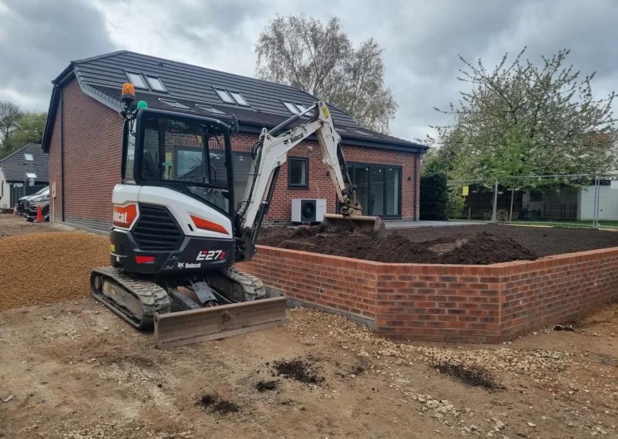 Tracked excavator working beside a newly built curved brick retaining wall during groundworks at a modern house