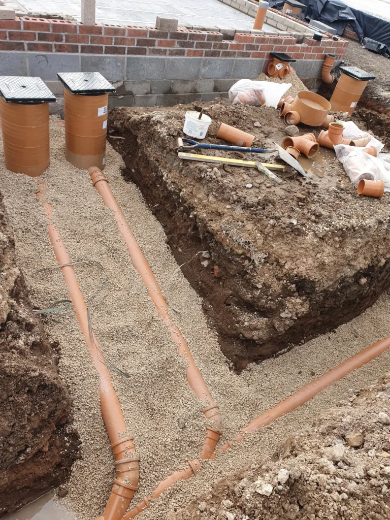 New orange foul drainage pipework and junctions bedded in pea gravel in an open trench beside a brick wall