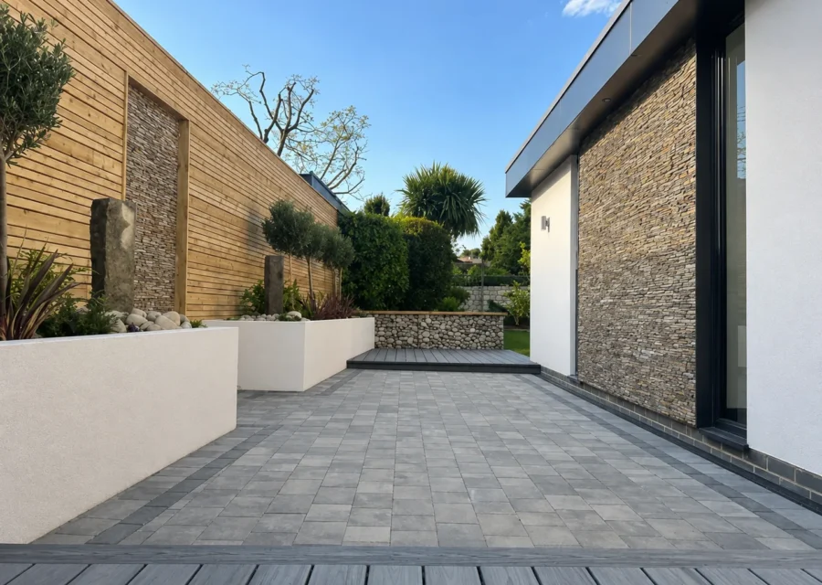 Grey block paved courtyard running between a cedar clad wall and a stone clad extension, with rendered planters, olive trees and a composite deck
